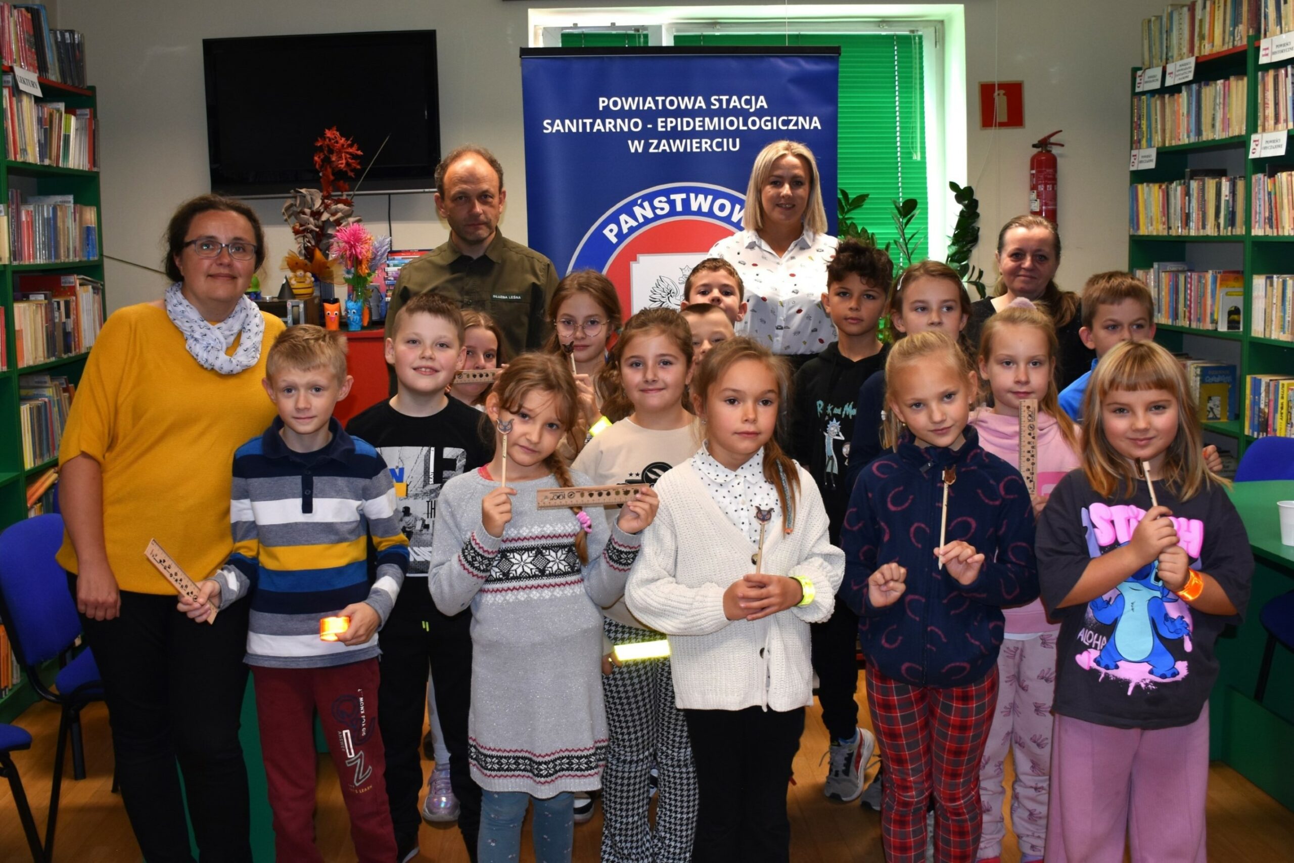 Grupa dzieci biorących udział w spotkaniu edukacyjnym o grzybach stojąca w bibliotece z leśniczym i pracownikiem sanepidu