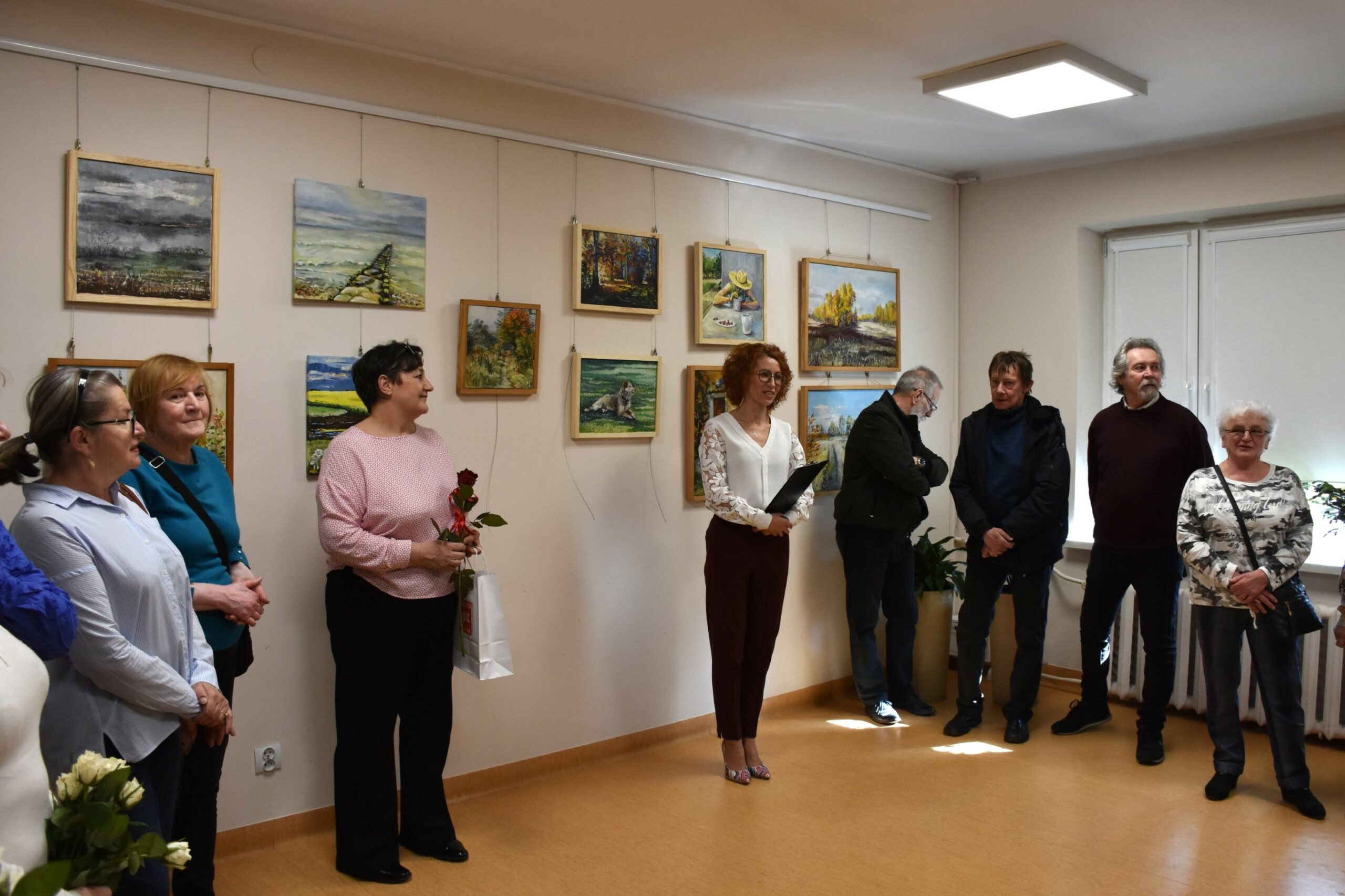 Dyrektor biblioteki przemawia do uczestników wernisażu ustawionych w półokręgu, wszyscy stoją, w tle obrazy olejne