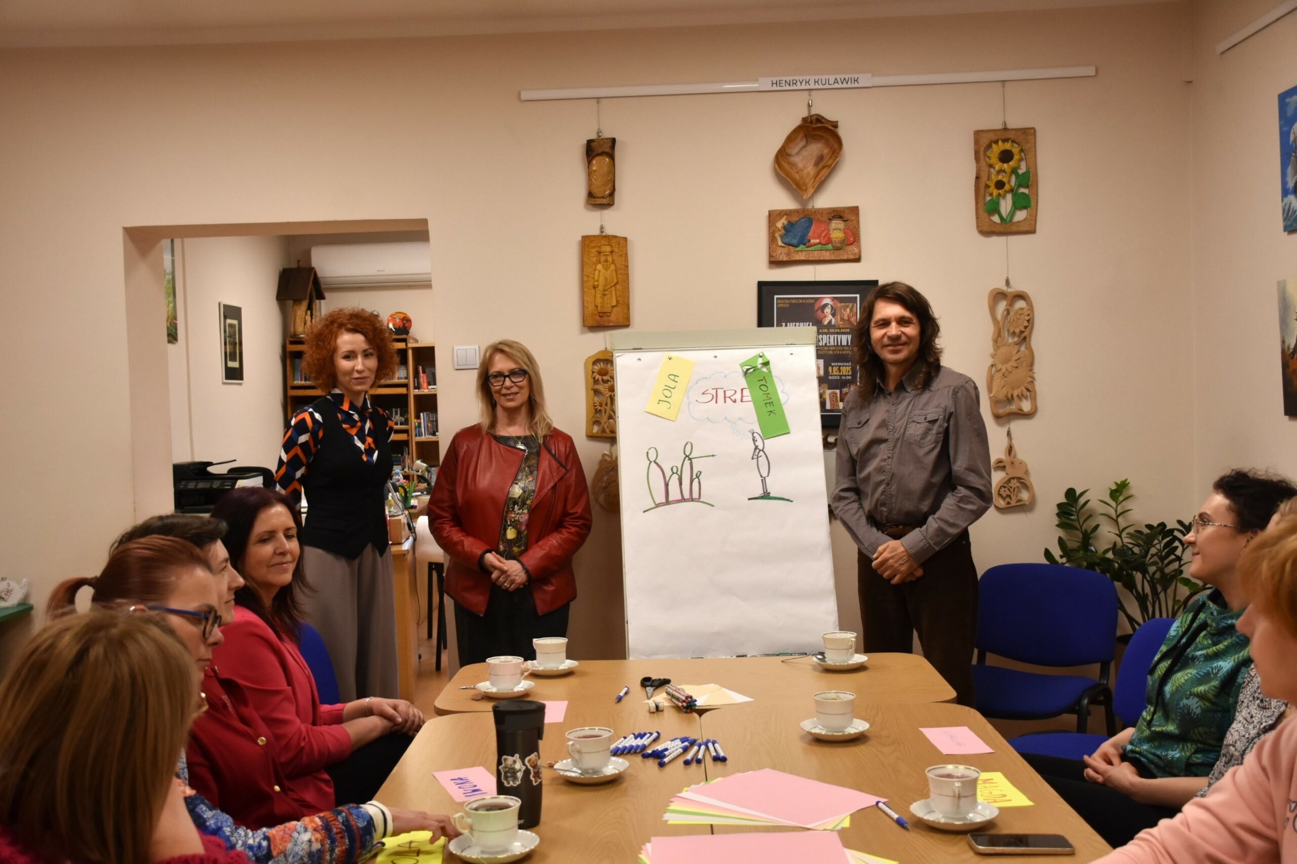 Uczestnicy warsztatu stres pod kontrolą siedzący przy stole, w tle stoi dwoje prowadzących i dyrektor biblioteki