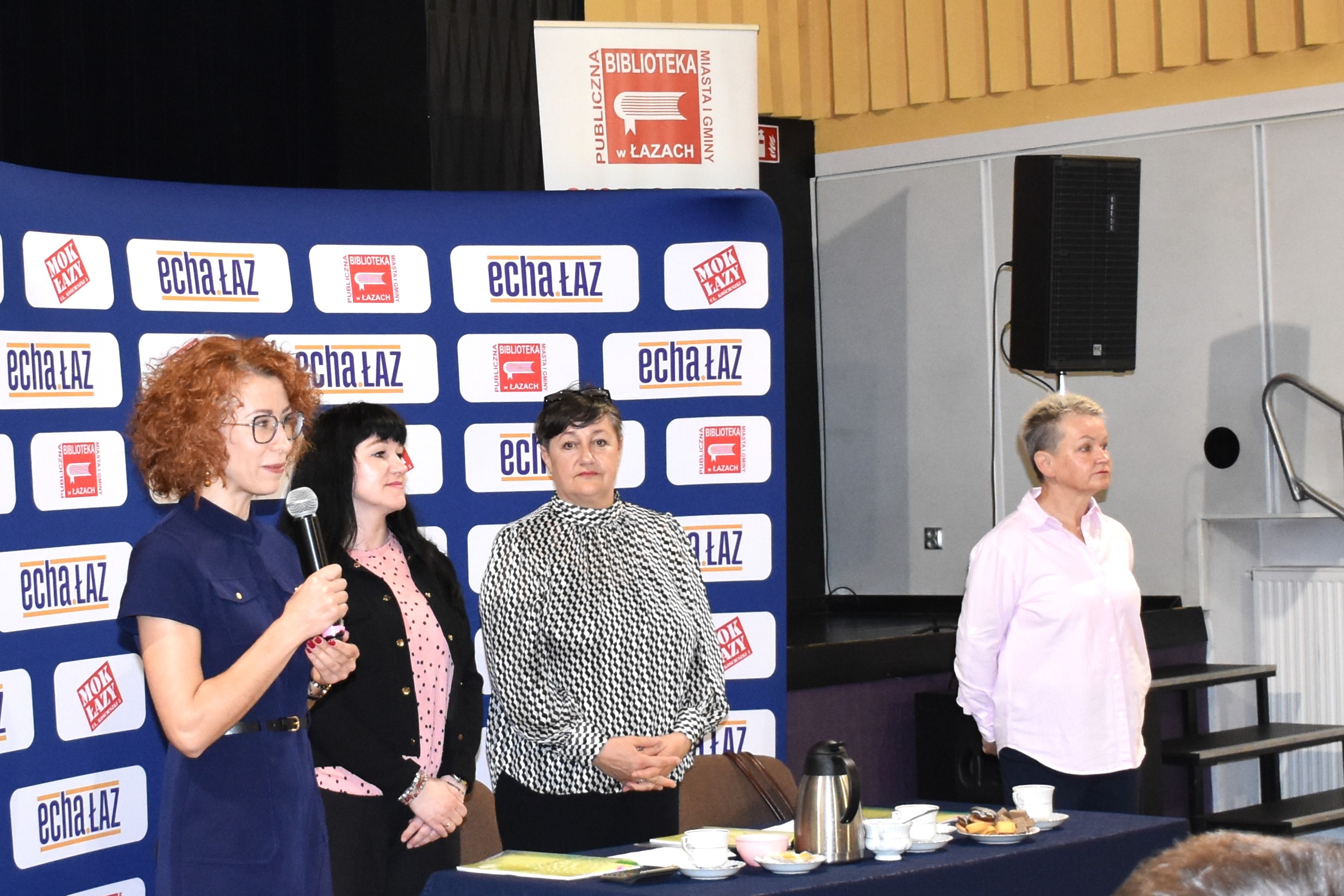 Dyrektor biblioteki mówiący do mikrofonu, w tle trzy kobiety - jury turnieju wiedzy o książce i bibliotece