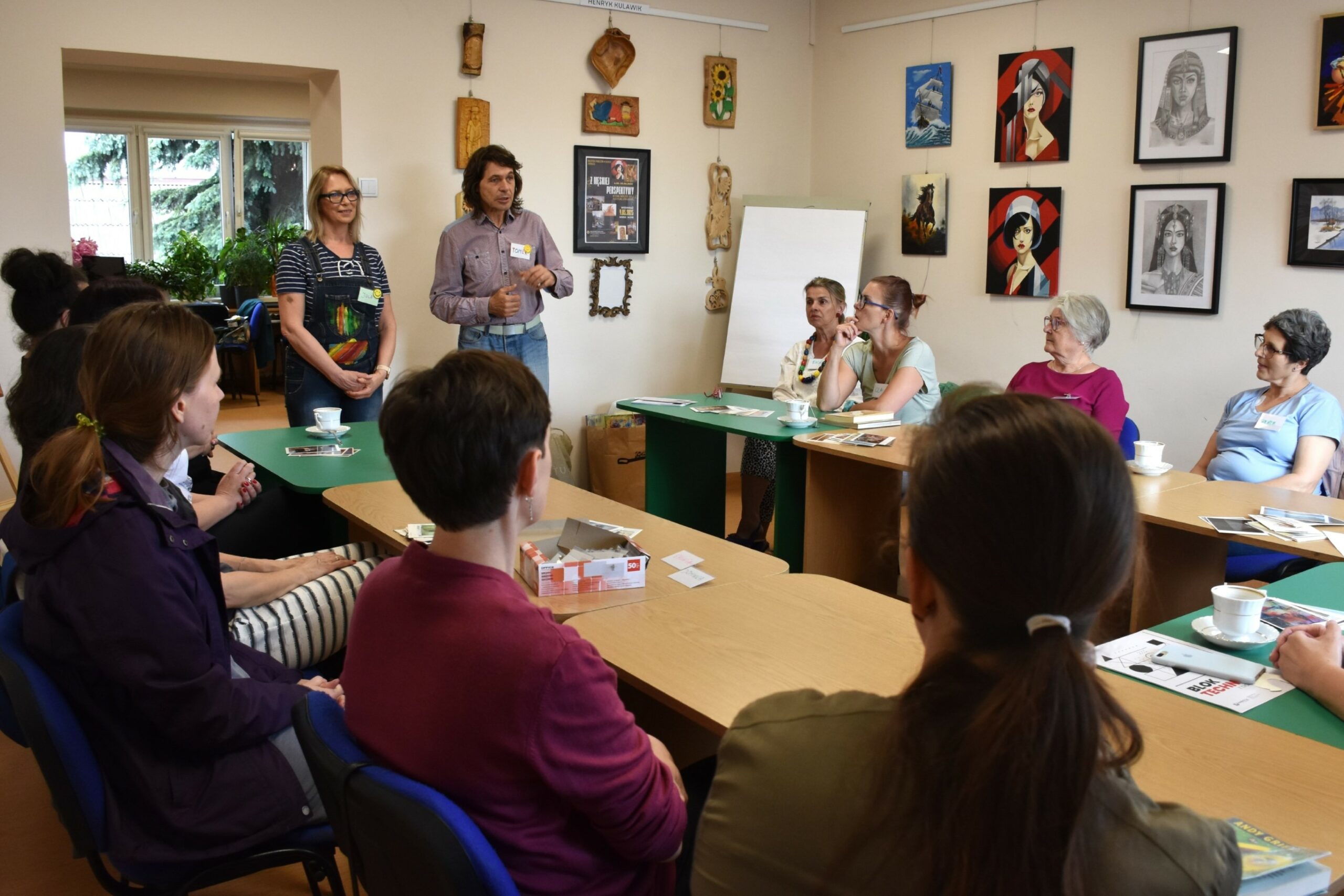 Grupa uczestniczek warsztatu paleta emocji ziedząca przy stołach, w tle prowadzący warsztat