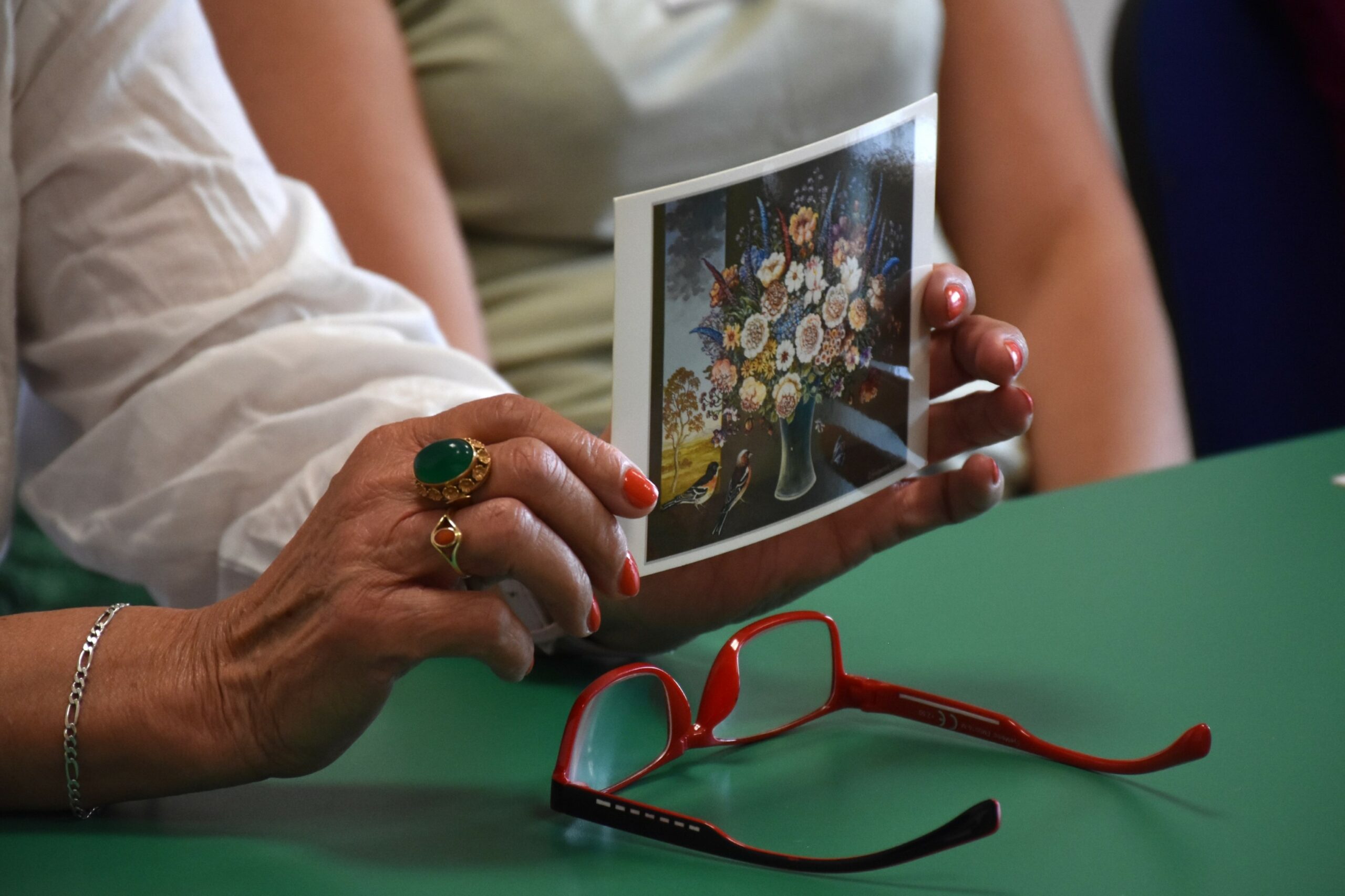 Pocztówka z reprodukcją obrazu kwiatów trzymana w rękach kobiety, z przodu okulary