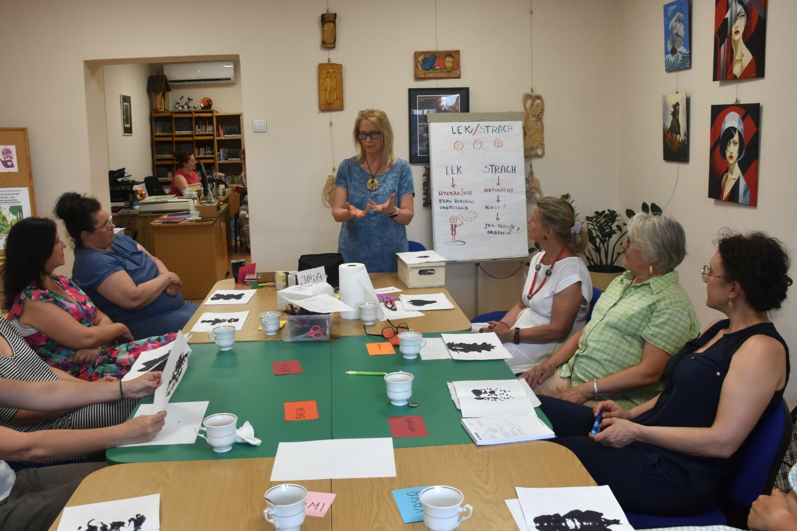 Prowadząca warsztat oswoić lęk przy flipcharcie, na pierwszym planie sześć kobiet siedzących za stołem i patrzących na prowadzącą