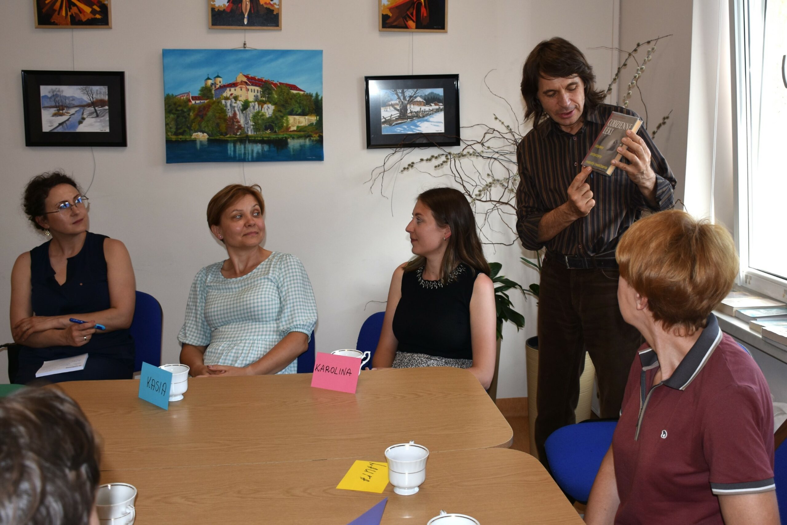 Prowadzący warsztat oswoić lęk prezentujący ksiażkę psychologiczną - w tle cztery kobiety przyglądające się mężczyźnie