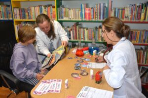 Dwie bibliotekarki przy stoliku z książkami, jedna z nich pokazuje kilka książek siedzącej przy stoliku nastolatce w lawendowej bluzie