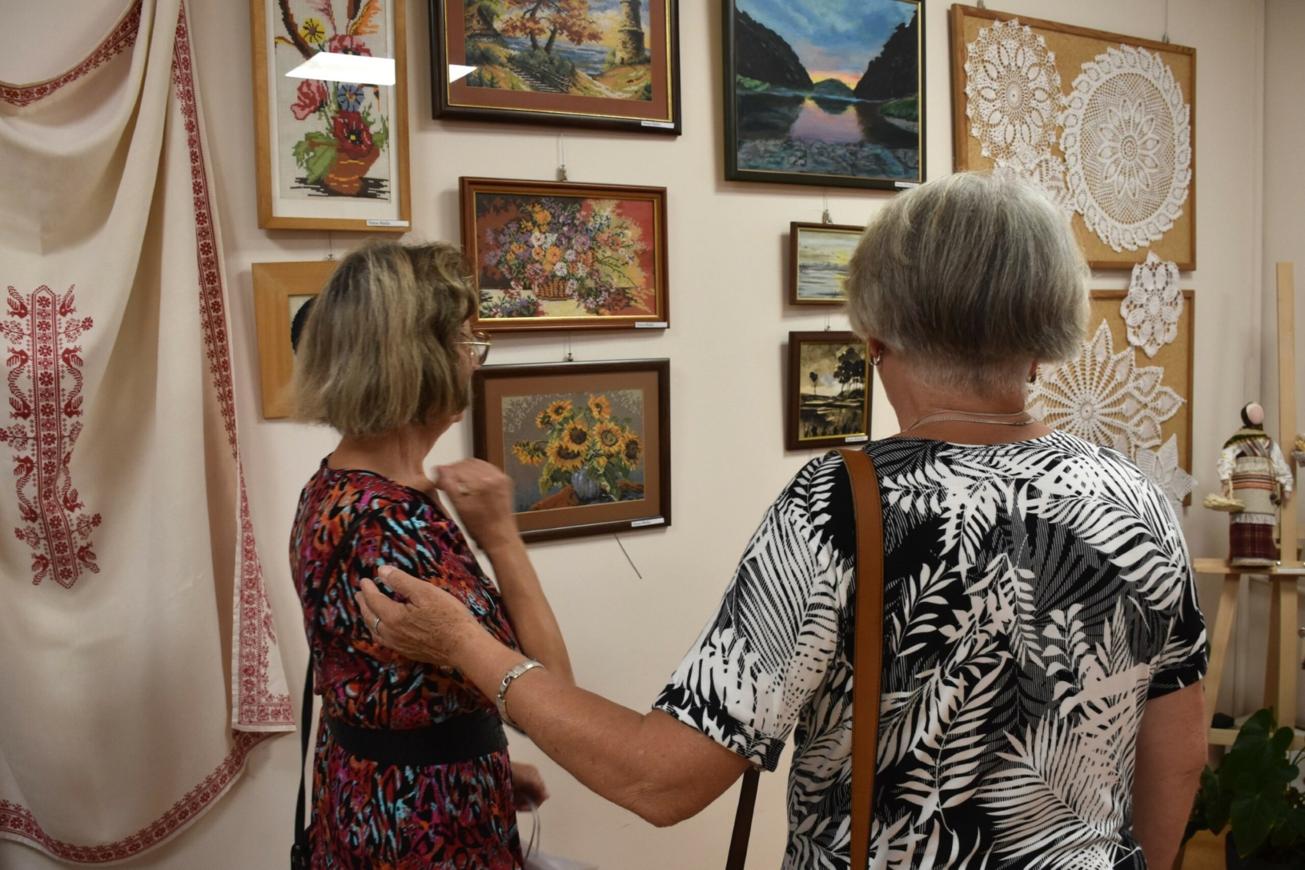Dwie seniorki wpatrzone w ścianę, na której wiszą obrazki haftowane krzyżykiem, zdjęcie przez plecy