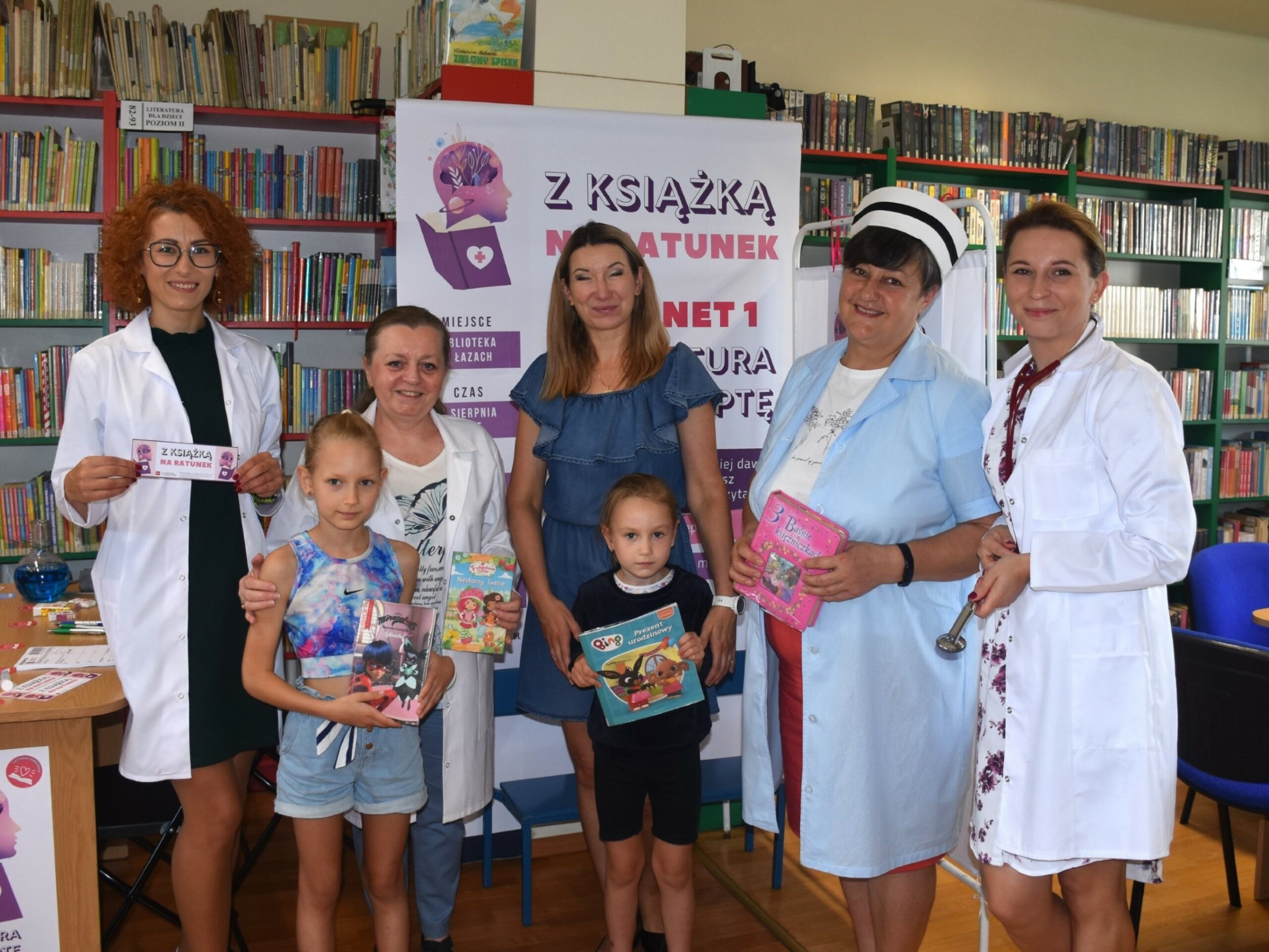 Mama, dwoje dzieci i cztery bibliotekarki w lekarskich kitlach, z książkami w ręku, w tle baner akcji Literatura na receptę