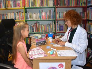 Nastolatka i bibliotekarka przebrana w lekarski kitel, siedzą przy stoliku, na stoliku zestaw książek, zakładki i menzurki z kolorową wodą