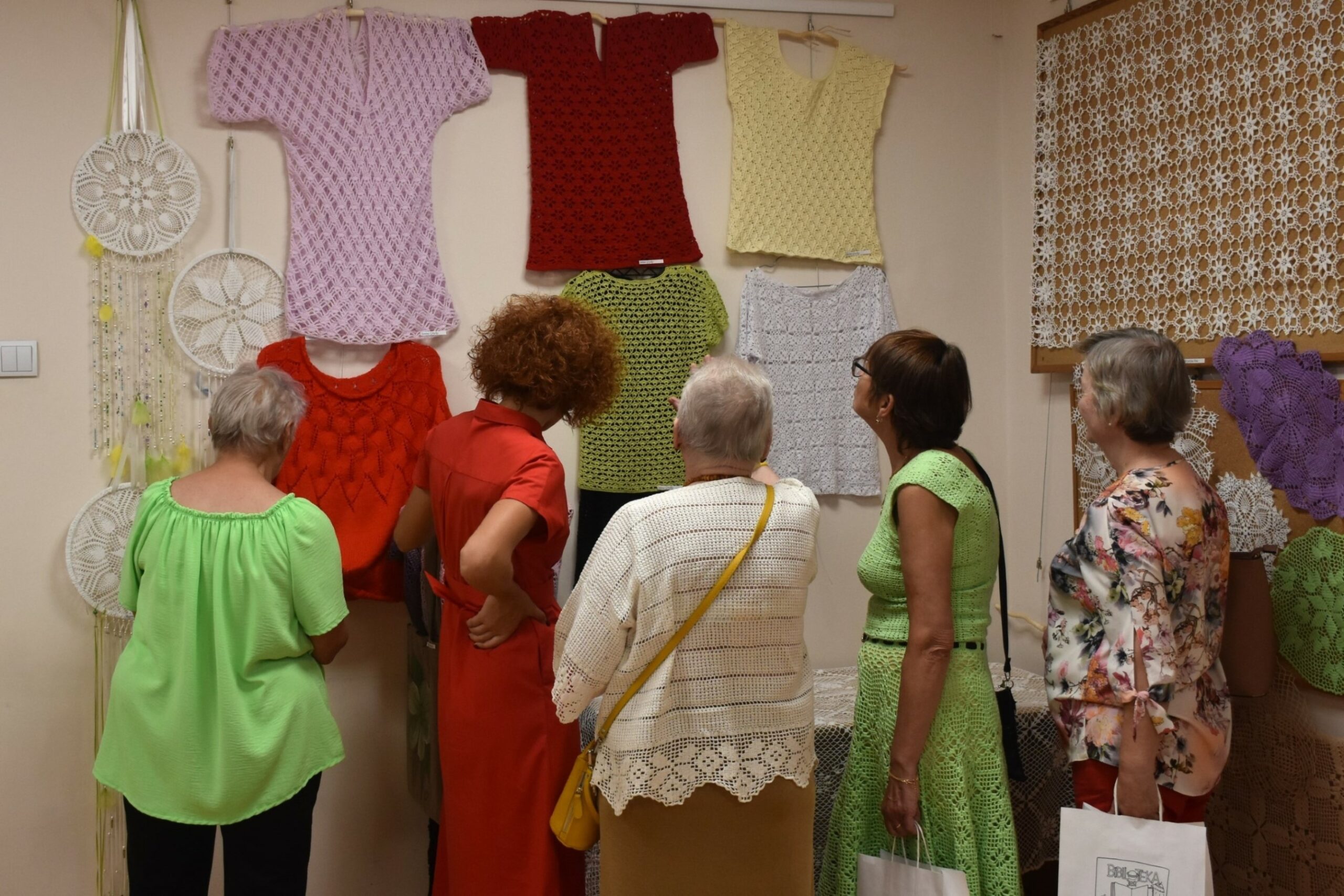 Pięć kobiet oglądających bluzki dziergane na szydełku wiszące na ścianie, zdjęcie przez plecy