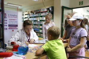 Przy stoliku bibliotekarka siedząca w lekarskim kitlu, wypisująca receptę, wokół niej czworo dzieci