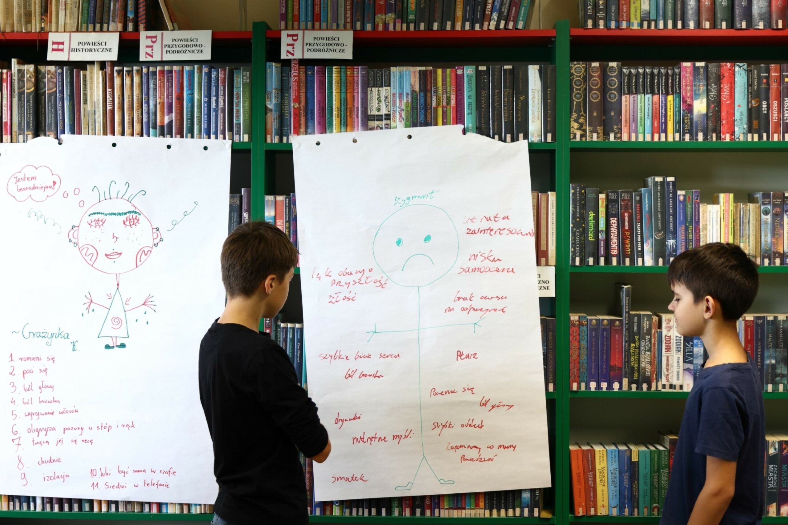 Dwoje chłopców patrzących na duży arkusz białego papieru, na którym narysowany jest schemat człowieka z oznaczeniem gdzie może w nim być stres