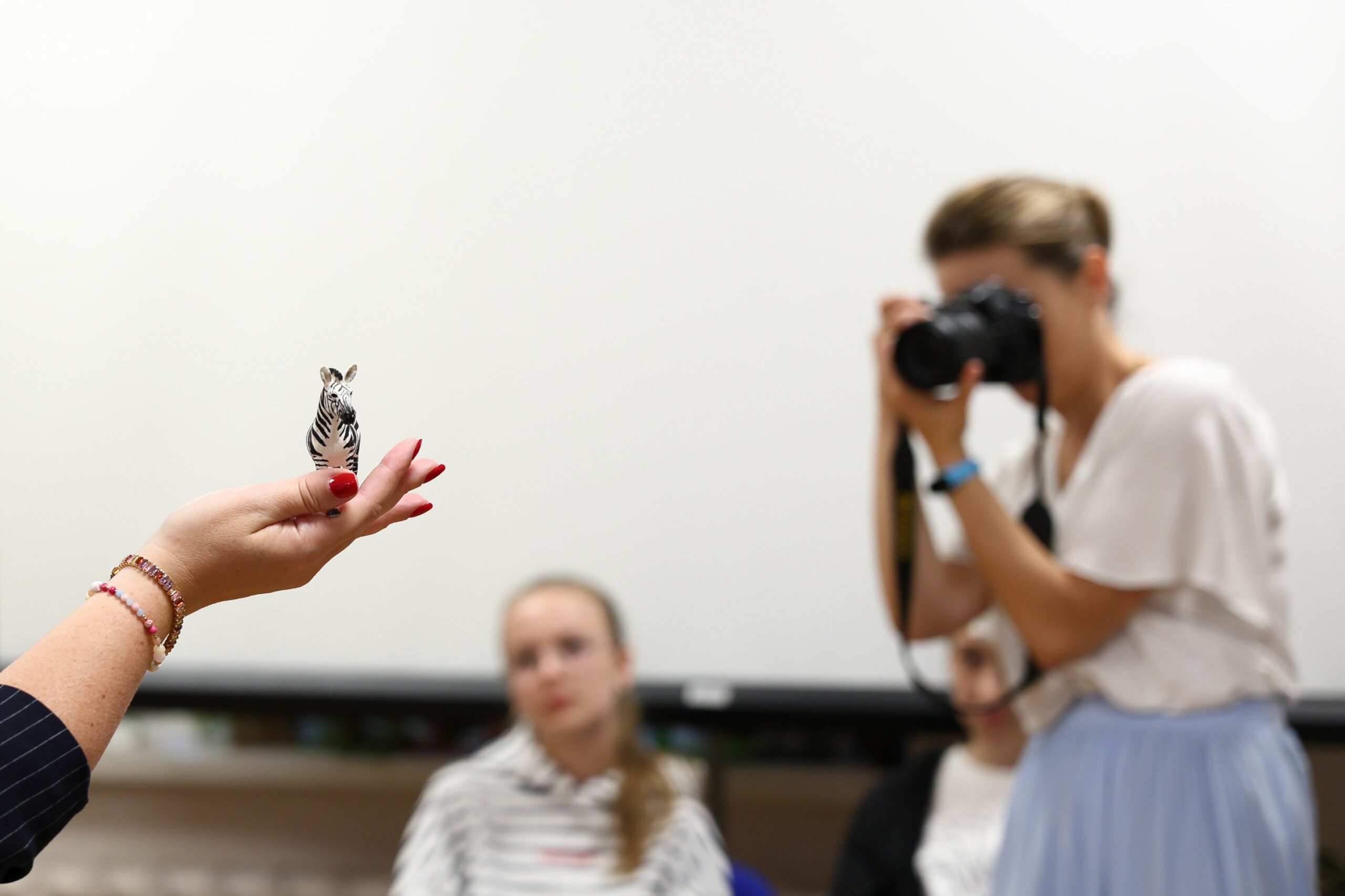 Na pierwszym planie kobieca dłoń, na której ustawiona jest figurka zebry, w tle rozmyty obraz kobiety z aparatem w ręku