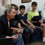 Siedząca na krześle kobieta z kokiem gestykulująca rękami, w tle czworo chłopców - nastolatków