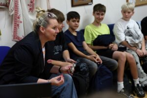 Siedząca na krześle kobieta z kokiem gestykulująca rękami, w tle czworo chłopców - nastolatków