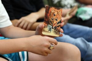 Zdjęcie przedstawiające kartę do gry trzymaną w chłopięcej ręce, na której narysowany jest trójoki potwór i mały człowiek wchodzący pod filiżankę