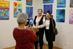 Dwie kobiety pozujące w uśmiechu na tle obrazów Jolanty Łysakowskiej, trzecia kobieta stojąca tyłem robi telefonem zdjęcie pozującym