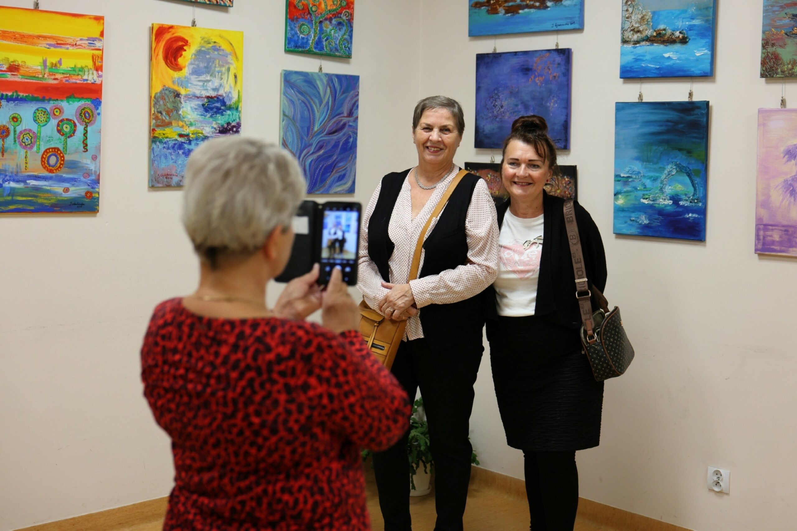Dwie kobiety pozujące w uśmiechu na tle obrazów Jolanty Łysakowskiej, trzecia kobieta stojąca tyłem robi telefonem zdjęcie pozującym