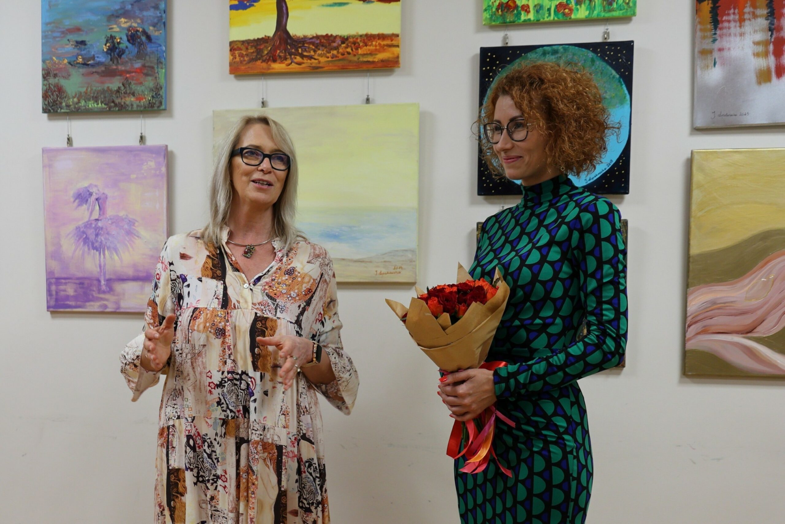 Jolanta Łysakowska i dyrektor biblioteki w Łazach stojące z uśmiechami na twarzy na tle obrazów autorki, dyrektor biblioteki trzyma w dłoniach bukiet czerwonych i pomarańczowych róż