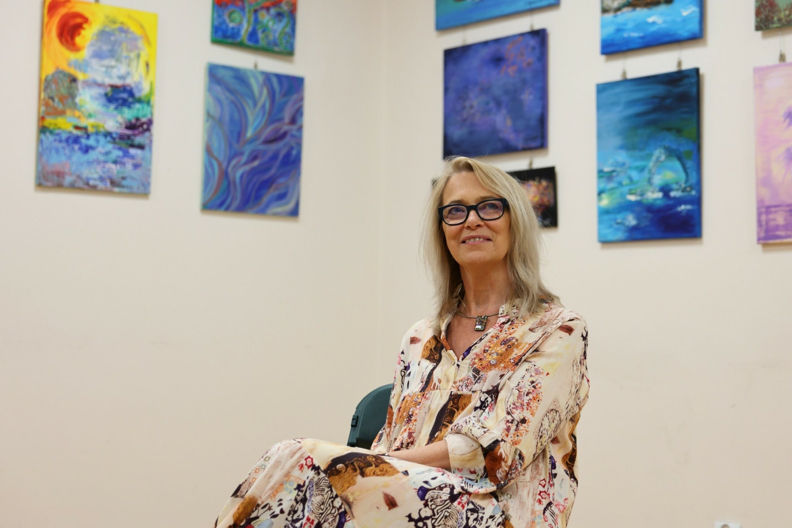 Jolanta Łysakowska w zamyśleniu, na drugim planie jej obrazy olejne