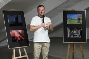 Autor fotografii z mikrofonem w ręku, po bokach sztalugi z jego pracami