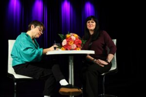 Autorka podpisująca książkę i kobieta czekająca na wpis, patrząca w obiektyw, siedzące przy stoliku, na którym leży duży bukiet kolorowych kwiatów