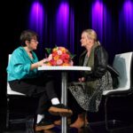 Autorka podpisująca książkę i kobieta czekająca na wpis, pogrążone w rozmowie, siedzące przy stoliku, na którym leży duży bukiet kolorowych kwiatów