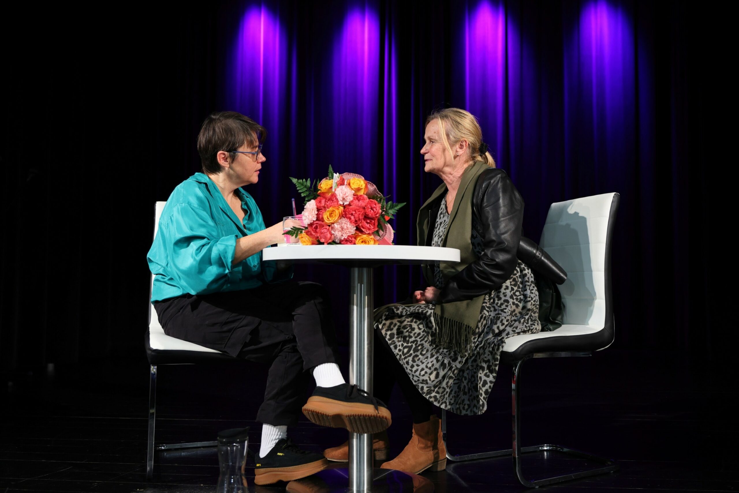 Autorka podpisująca książkę i kobieta czekająca na wpis, pogrążone w rozmowie, siedzące przy stoliku, na którym leży duży bukiet kolorowych kwiatów