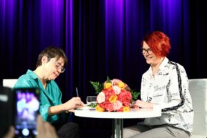 Autorka podpisująca książkę i kobieta czekająca na wpis, siedzące przy stoliku, na którym leży duży bukiet kolorowych kwiatów