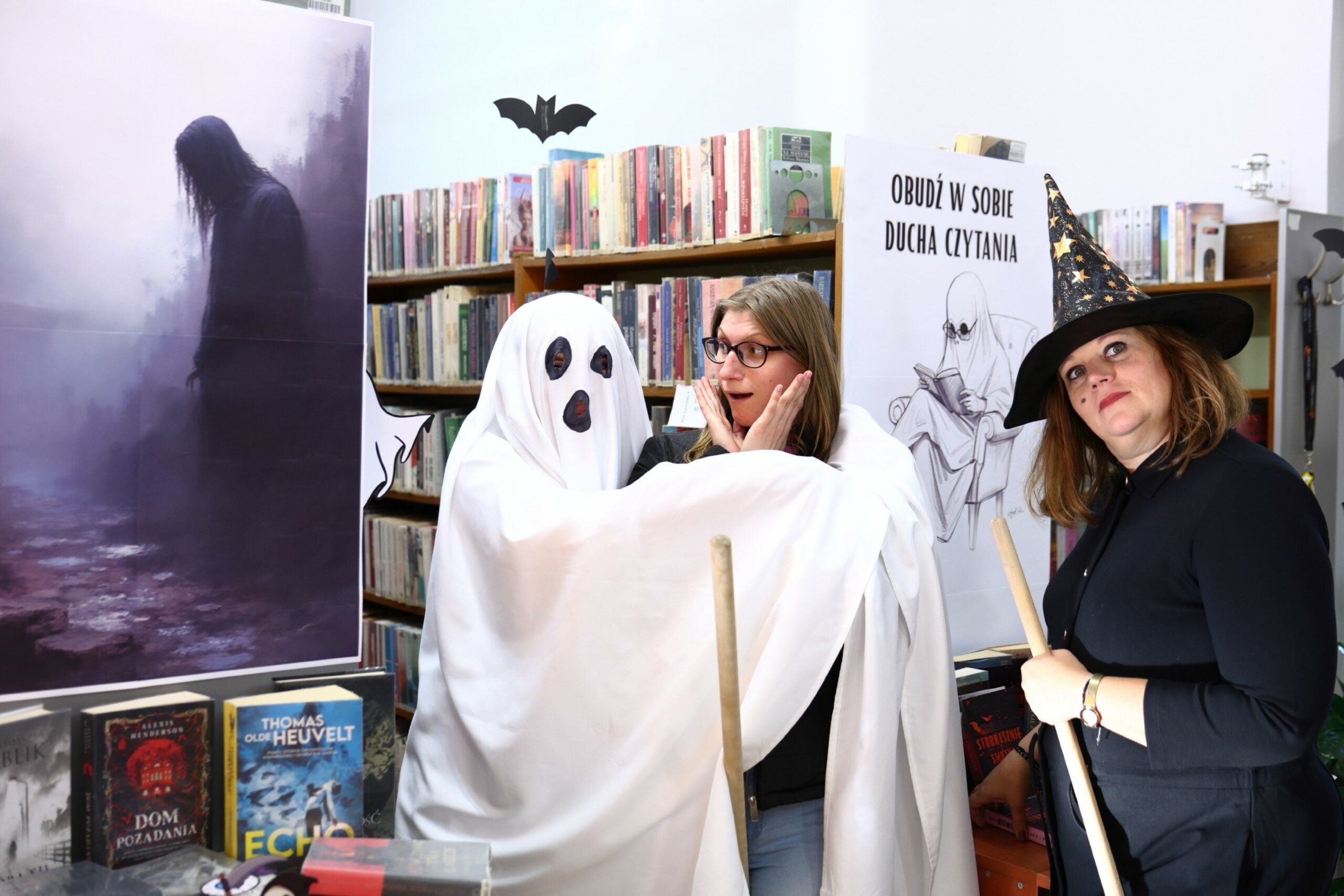 Czytelniczka z przerażona miną w objęciach ducha, po lewej stronie wpatrująca się w obiektyw czarownica