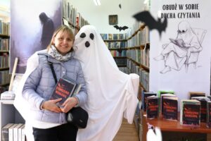 Duch i czytelniczka z ksiażką w ręku pozujące na tle regałów z książkami, z sufitu zwisają papierowe nietoperze