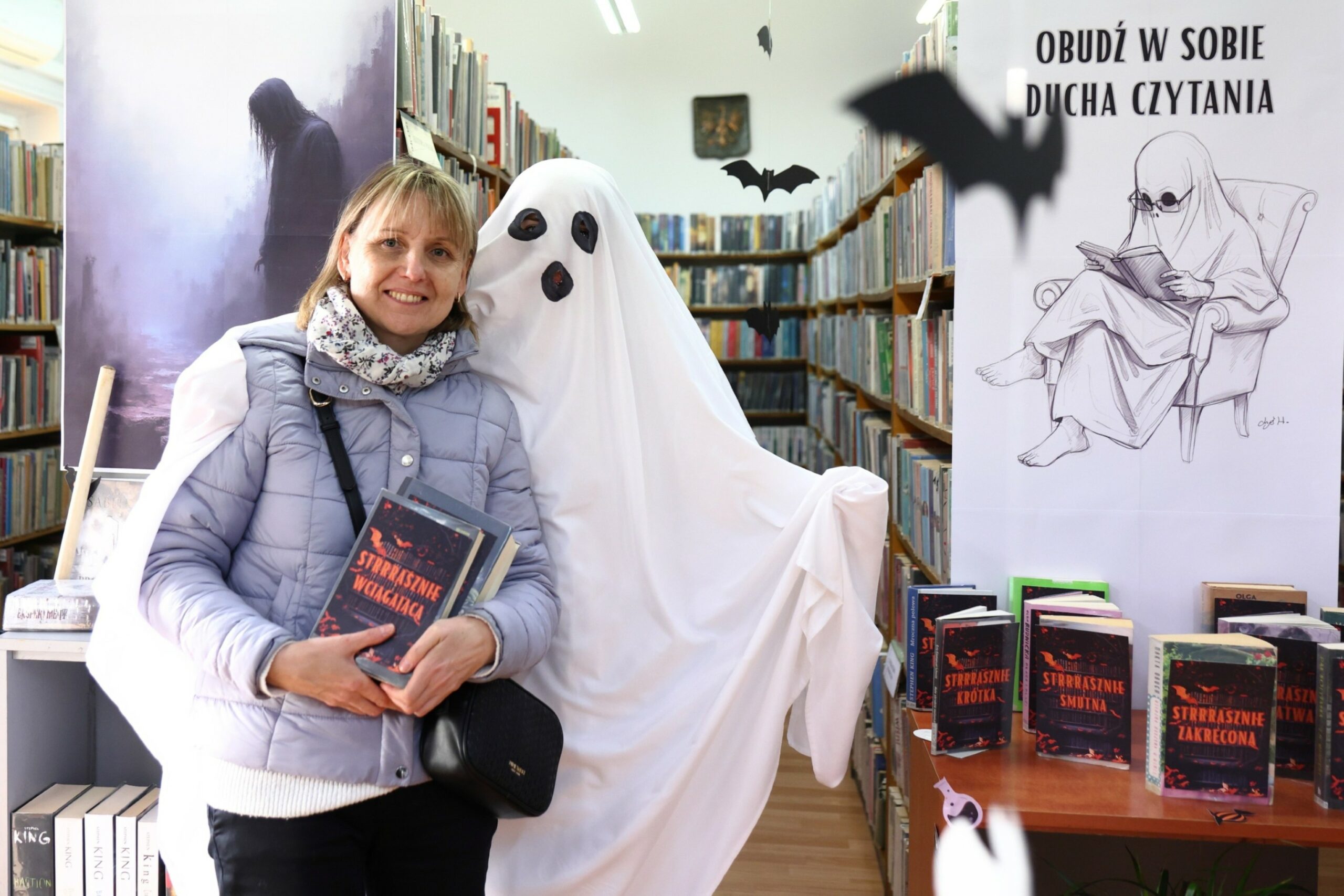 Duch i czytelniczka z ksiażką w ręku pozujące na tle regałów z książkami, z sufitu zwisają papierowe nietoperze