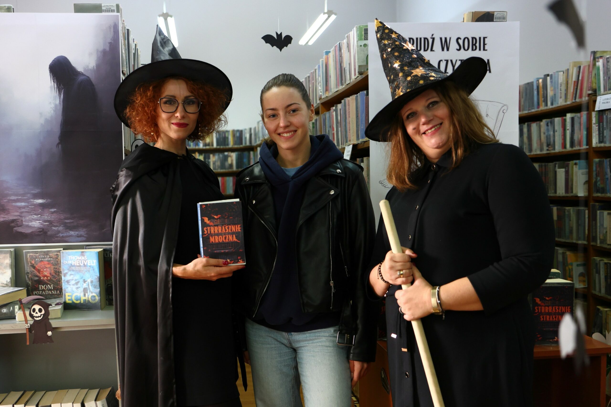 Dwie bibliotekarki w strojach czarownic i czytelniczka na tle regałów z książkami