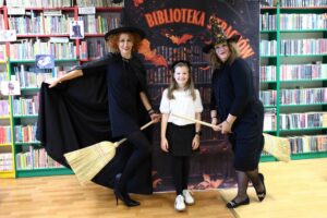 Dwie bibliotekarki w strojach czarownic i dziewczynka biorąca udział w akcji Biblioteka strachów pozują na tle książek