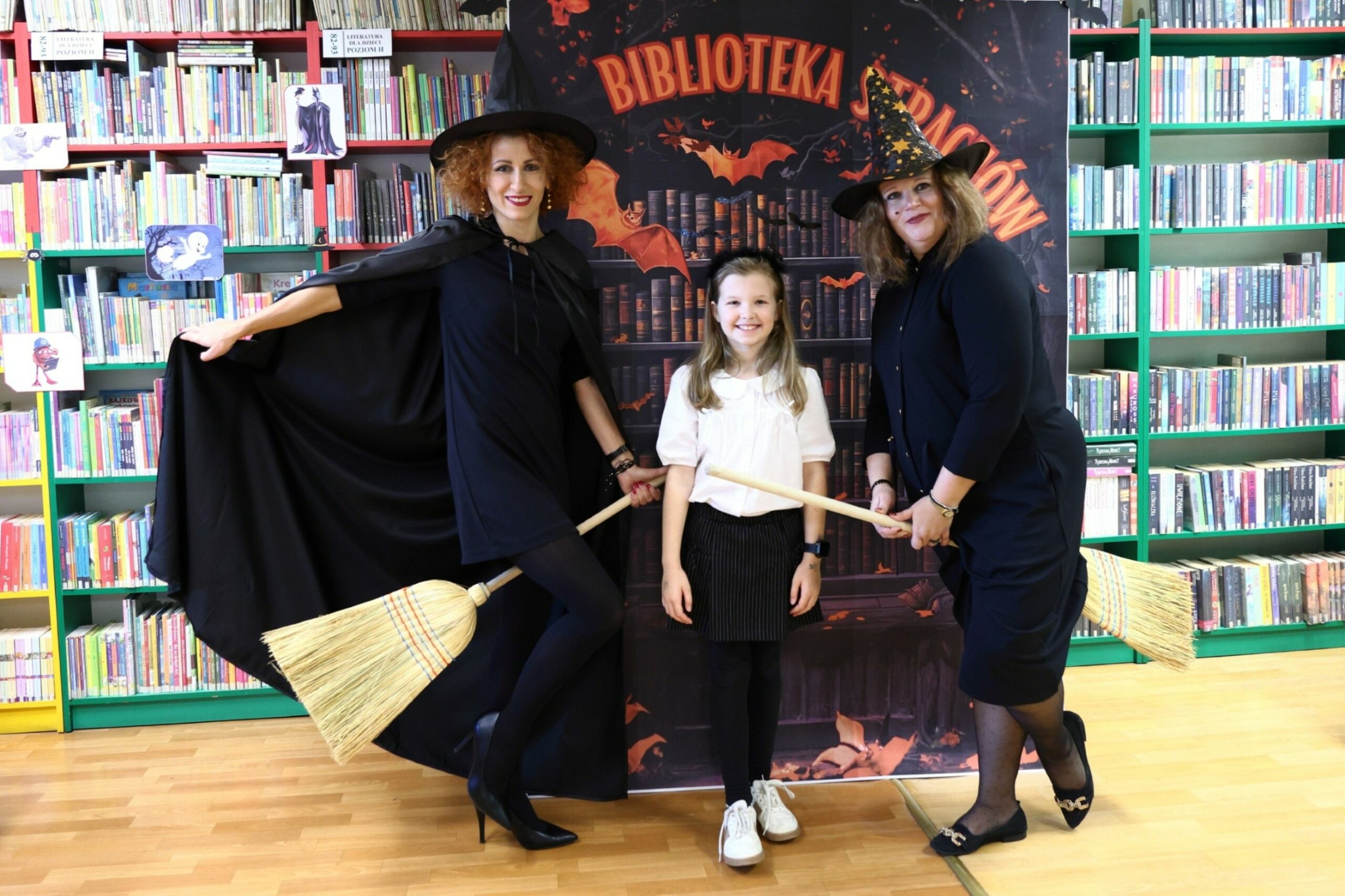Dwie bibliotekarki w strojach czarownic i dziewczynka biorąca udział w akcji Biblioteka strachów pozują na tle książek