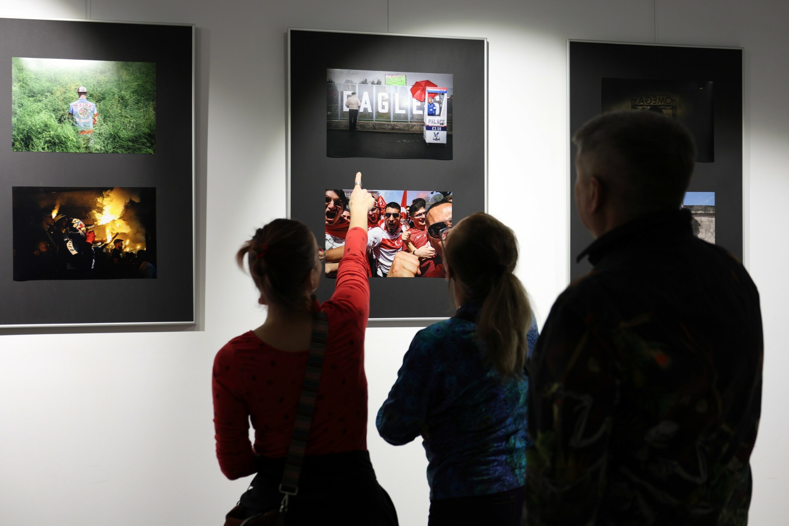 Trzy osoby tyłem, wpatrujące się w ścianę z fotografiami w antyramach, kobieta z lewej strony wskazuje palcem jedną z nich