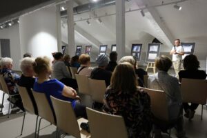 Widownia wernisażu na siedząco, wpatrująca się w autora fotografii otoczonego antyramami na sztalugach, widok zza jej pleców