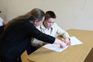 Chłopiec siedzący przy biurku, wpatrzony w kartę testu i bibliotekarka wskazująca na jedno z pytań na karcie