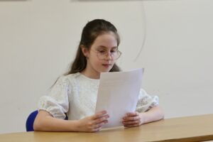 Uczennica siedząca przy stoliku i czytająca tekst z białej kartki