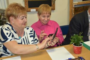 Dwie kobiety w uśmiechu - uczestniczki warsztatu Internet pod lupą, jednaq z nich trzyma w ręku telefon i pokazująca na nim coś drugiej uczestniczce