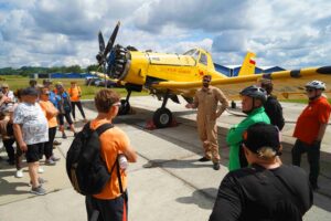 Pilot stojący przy żółym samolocie dromader otoczony kilkoma ludźmi