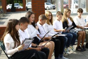 Grupa ośmiu nastolatków siedzących na krzesłach w przestrzeni miejskiej, trzymających w rękach teksty, w tle woda fontanny