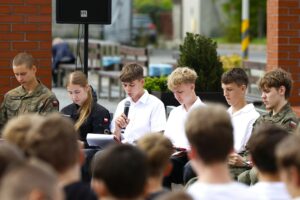 Grupa sześciu nastolatków siedzących na krzesłach w przestrzeni miejskiej, w tym jeden mówiący do mikrofonu, widok zza pleców widowni