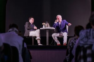 Autor i dziennikarz prowadzący spotkanie Granice świata siedzący przy stoliku na scenie miejskiego ośrodka kultury - widok z perspektywy końca sali